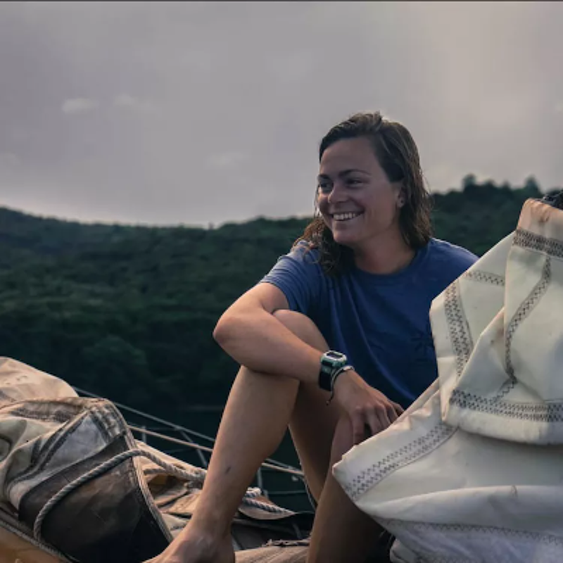 The image shows a woman on a boat, smiling. She is wearing a blue shirt and a watch. The background features a cloudy sky and a green, forested hill. The overall mood is peaceful and serene, suggesting a moment of enjoyment and relaxation on the water.